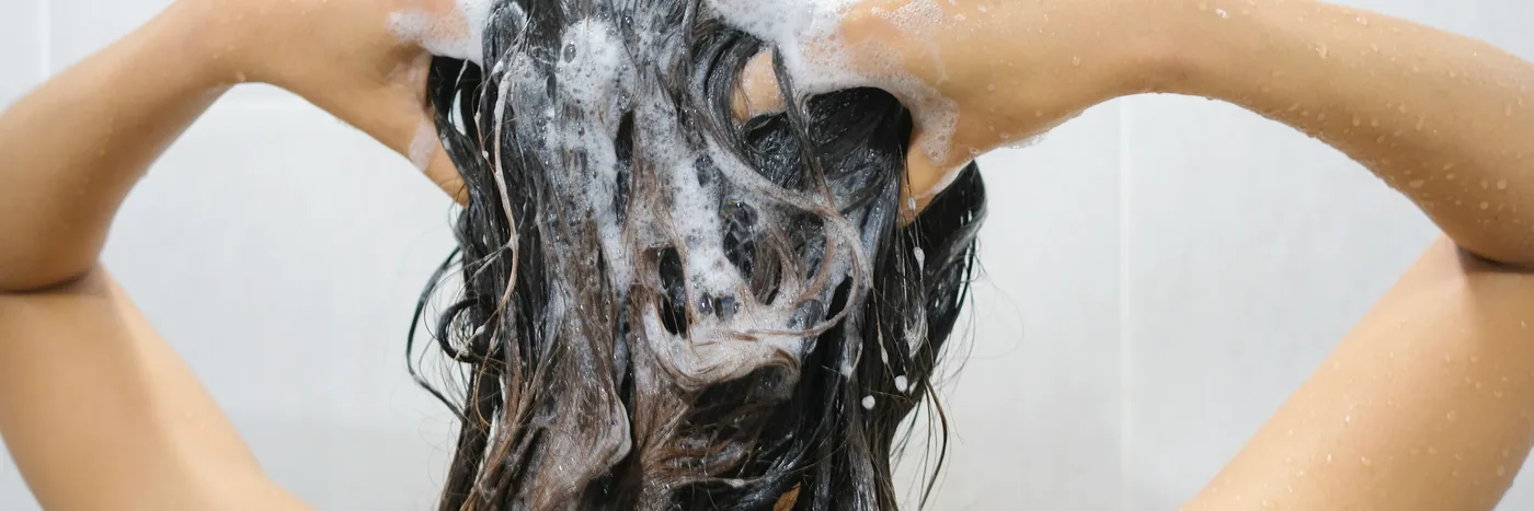 A woman working shampoo lather through long dark hair with both hands under a shower.