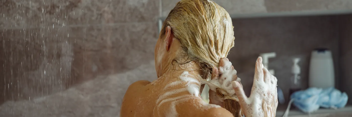 A woman seen from behind washing lathered hair under a shower, shampoo bottles on the shelf behind her.