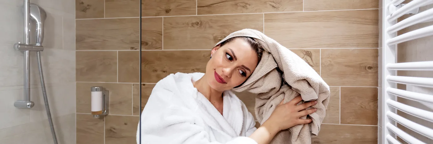 A woman in a white robe drying her hair with a towel in a tiled bathroom after a shower.